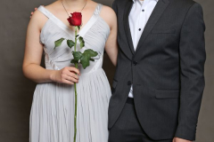 Paar steht vor grauem Hintergrund; Frau im hellen Kleid hält eine rote Rose, Mann im dunklen Anzug.