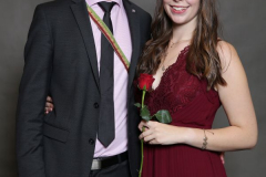 Mann im Anzug und Frau im bordeauxroten Kleid stehen nebeneinander; sie hält eine rote Rose.