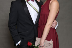 Mann im Anzug und Frau im burgunderfarbenen Kleid stehen nebeneinander; sie hält eine Clutch und eine rote Rose.