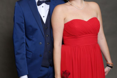 Mann im blauen Anzug und Frau im roten trägerlosen Kleid, beide vor grauem Hintergrund; sie hält eine rote Rose.