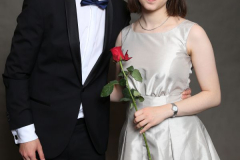 Mann im schwarzen Anzug und Frau im hellen Kleid mit roter Rose, vor grauem Hintergrund.