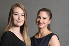 Zwei Frauen in schwarzen Kleidern stehen nebeneinander vor grauem Hintergrund.
