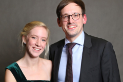 Frau im grünen Abendkleid und Mann im dunklen Anzug stehen nebeneinander vor grauem Hintergrund.