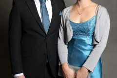 Mann im schwarzen Anzug und Frau im blauen Kleid stehen nebeneinander vor grauem Hintergrund.