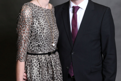 Zwei Personen stehen vor grauem Hintergrund; links trägt eine ein gemustertes Kleid, rechts ein Mann einen Anzug.