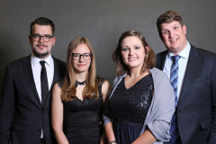 Vier Menschen in festlicher Kleidung vor grauem Hintergrund.