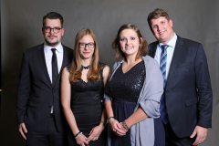Vier Personen in eleganter Kleidung vor dunklem Hintergrund, zwei Männer im Anzug und zwei Frauen in Kleidern.