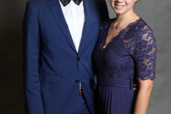 Mann im blauen Anzug mit Fliege und Frau im blauen Spitzenkleid stehen vor grauem Hintergrund.