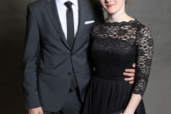 Mann im dunklen Anzug und Frau im schwarzen Spitzenkleid stehen nebeneinander vor grauem Hintergrund.