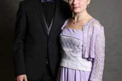Mann im schwarzen Anzug und Frau in violettem Kleid stehen vor grauem Hintergrund.