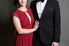 Frau in rotem Abendkleid und Mann im schwarzen Anzug stehen vor grauem Hintergrund.