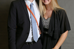 Ein Mann im Anzug und eine Frau im schwarzen Kleid stehen nebeneinander vor grauem Hintergrund.