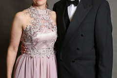 Frau im rosa Spitzenkleid und Mann im schwarzen Smoking stehen nebeneinander vor grauem Hintergrund.
