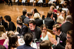 Großer Saal mit vielen sitzenden Personen an langen Tischen, Gläsern und weißen Tischdecken.