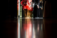 Tänzergruppe in Kostümen steht in einer Türöffnung; dunkler Raum, Spiegelung auf dem Boden.