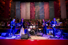 Band auf einer Bühne mit Sängerin, Keyboard, Schlagzeug und zwei Gitarren; im Hintergrund Tische und Banner.