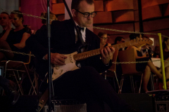 Mann spielt E-Gitarre auf einer Bühne vor Mikrofonständer und Lautsprecher, im Hintergrund sitzende Personen.