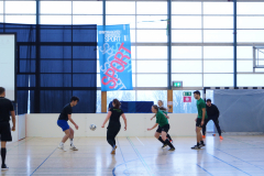 Mehrere Personen spielen in einer Halle Fußball, ein Ball liegt am Boden; im Hintergrund ein blaues Banner.