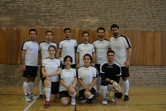 Elf Personen in weißen und schwarzen Sportshirts stehen und knien vor einer Backstein- und Holzwand.