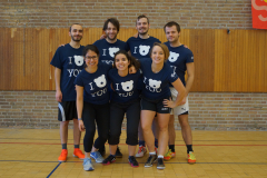 Sieben Personen in blauen T-Shirts stehen in einer Turnhalle vor einer Holz- und Backsteinwand.