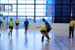 Mehrere Personen spielen in einer Sporthalle Fußball; ein Tor, ein Banner und große Fenster sind zu sehen.