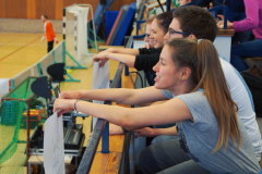 Mehrere Personen sitzen an einer Hallenbrüstung und halten weiße Blätter; im Hintergrund ein Sportfeld.