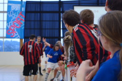 Hallenszene mit mehreren Personen, teils in rot-schwarzen und blauen Sporttrikots, neben einem Banner mit „SPORT“.