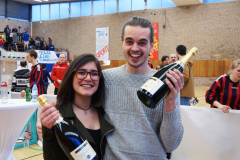 Zwei lächelnde Personen halten jeweils eine Sektflasche in einer Sporthalle.