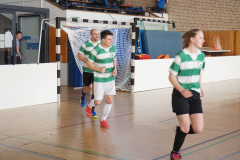 Drei Spieler in grün-weiß gestreiften Trikots laufen in einer Sporthalle vor einem Handballtor.