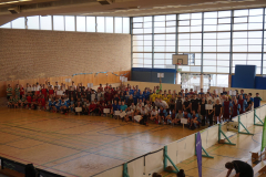 Große Gruppe von Personen in Sportkleidung in einer Turnhalle, einige halten Schilder; Basketballkorb im Hintergrund.
