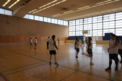 Mehrere Personen in Sportkleidung in einer Turnhalle, Basketballkorb an der Wand, große Fenster und Holzfußboden.