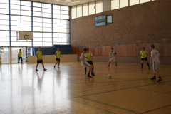 Mannschaft in einer Sporthalle spielt Fußball; mehrere Spieler, Tor links, Ball am Boden, Anzeigetafel an der Wand.
