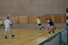 Mehrere Personen spielen Hallenfußball in einer Turnhalle mit Holzwand und Brickwand.