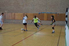 Vier Personen spielen Hallenfußball in einer Sporthalle; rechts steht eine Torhüterin vor dem Tor.