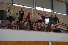 Mehrere Personen sitzen und stehen auf einer Tribüne hinter blauen Geländern in einer Halle.