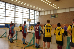 Mehrere Männer stehen in einer Sporthalle an einer Bande; im Hintergrund ein Spielfeld, Tore und eine Anzeigetafel.