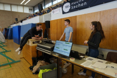 Drei Personen in einer Sporthalle an einem Tisch mit Monitor, Papierstapeln und Technik; im Hintergrund Zuschauer auf der Empore.
