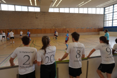 Indoor-Floorballspiel in einer Halle, Zuschauer im Vordergrund mit weißen Trikots an der Bande.
