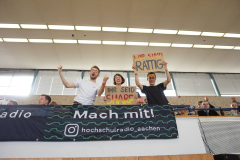 Drei Personen in einer Tribüne halten Protestschilder; unten hängt ein Banner mit „Mach mit!“ und „hochschulradio_aachen“.