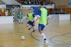 Hallensport mit drei Spielern in grünen Westen, Ball am Boden, Tor und Zuschauer auf der Tribüne.
