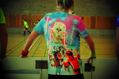 Person von hinten in buntem T-Shirt lehnt an einer Hallenbande; im Hintergrund laufen mehrere Personen in einer Sporthalle.