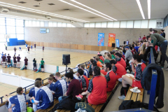 Menschen in einer Sporthalle auf Tribünen, auf dem Spielfeld mehrere Spieler, an der Wand Banner und ein Basketballkorb.