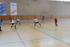Hallenszene mit mehreren Personen beim Fußballspiel; ein Ball liegt vor einer Spielerin in Weiß.