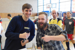 Zwei Männer mit Mikrophonen in einer Sporthalle; im Hintergrund stehen mehrere Personen, Basketballkorb und Fenster.