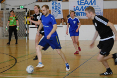 Mehrere Spieler in blau und schwarz spielen Hallenfußball; links steht ein Torwart vor dem Tor.