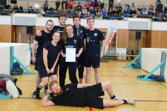 Sechs Personen in schwarzer Sportkleidung in einer Turnhalle; eine hält ein Blatt, eine liegt vorne.