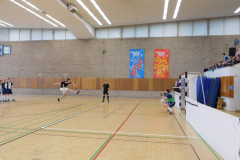 Sporthalle mit Handballspiel: Spieler springt, Torwart hält den Ball, Schiedsrichter in der Mitte.