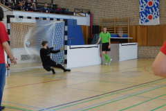 Handballtor in Halle, Torwart im Sprung, Ball im Netz, Spieler in Grün und Zuschauer auf der Tribüne.