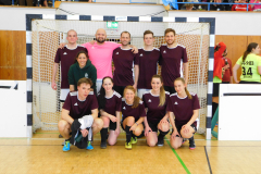 Elf Personen in Sportkleidung posieren vor einem Handballtor in einer Sporthalle.