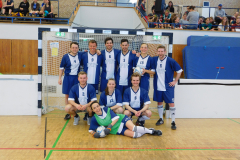 Mannschaft in blauen Trikots vor dem Tor in einer Sporthalle, Zuschauer auf der Tribüne.